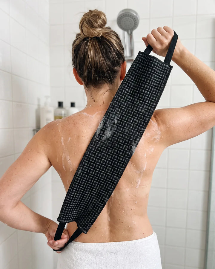 Photo d'exemple : femme de dos sous la douche qui se frotte le dos avec la serviette exfoliante noire tenue par ses deux poignées