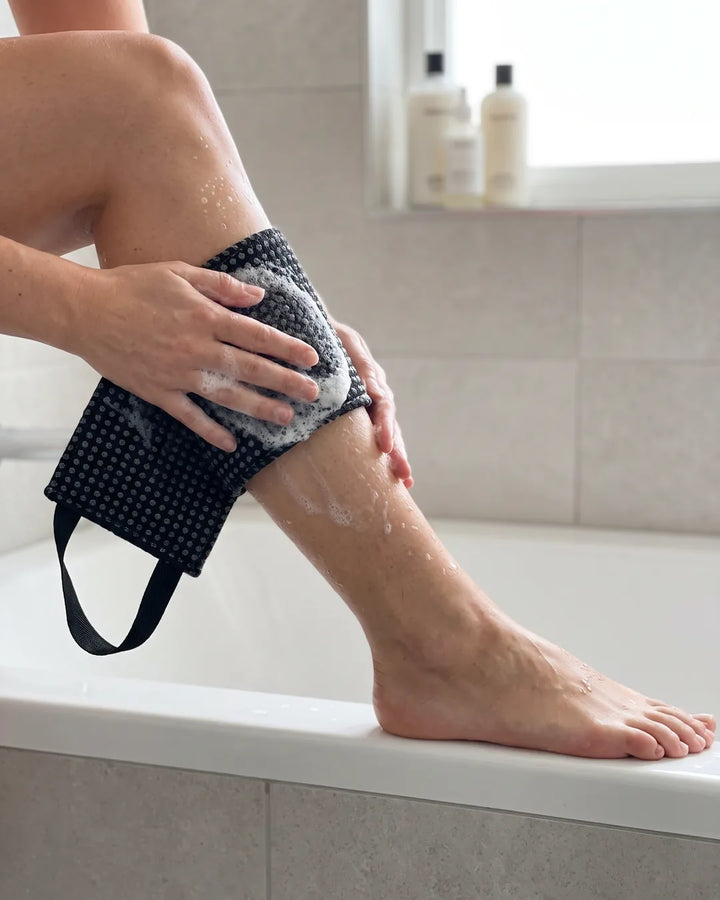 Photo d'exemple : mollet savonné frotté avec la serviette exfoliante noire, pied posé sur le bord de la baignoire